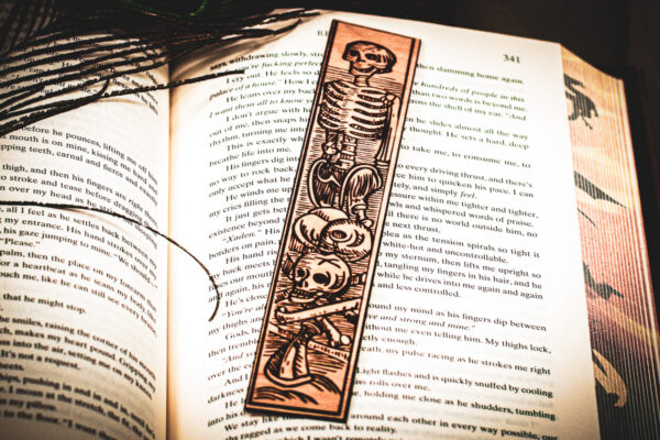A laser cut and engraved bookmark featuring a scene of skeletons and bones in a medieval woodcut style on the page of an open book.