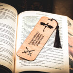 A laser cut and engraved bookmark with the quote, "A dragon without a rider is a tragedy. A rider without a dragon is dead." with two flying dragons at the top and two crossed swords cut out at the bottom with a burgundy tassel on top of the page of an open book.
