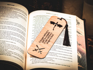 A laser cut and engraved bookmark with the quote, "A dragon without a rider is a tragedy. A rider without a dragon is dead." with two flying dragons at the top and two crossed swords cut out at the bottom with a burgundy tassel on top of the page of an open book.