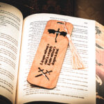 Handcrafted wooden bookmark with a cream tassel, featuring an engraved dragon silhouette and crossed swords, resting vertically on a book page.