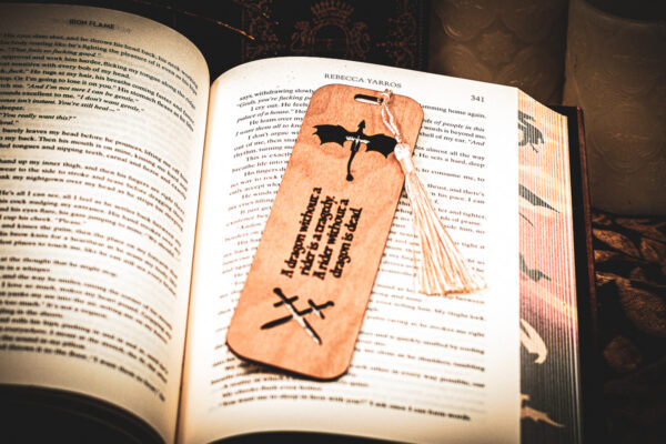 Handcrafted wooden bookmark with a cream tassel, featuring an engraved dragon silhouette and crossed swords, resting vertically on a book page.
