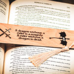 Detailed close-up of a laser-cut dragon bookmark with a cream-colored tassel, highlighting the sharp engraving of the dragon rider quote and intricate sword iconography.