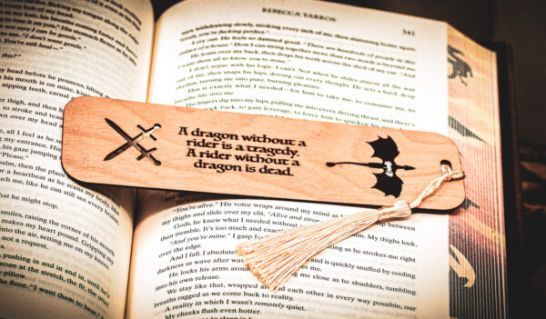 Detailed close-up of a laser-cut dragon bookmark with a cream-colored tassel, highlighting the sharp engraving of the dragon rider quote and intricate sword iconography.
