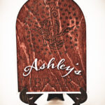 The front of a laser cut and engraved sign made of 1/8" birch plywood stained dark walnut engraved A wooden arched library sign from The Griffin and Stag featuring an engraved dragon within a circle of stars, with "Ashley's Library" in white script and serif lettering.