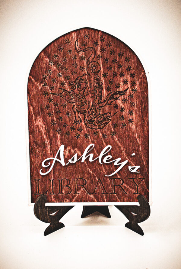 The front of a laser cut and engraved sign made of 1/8" birch plywood stained dark walnut engraved A wooden arched library sign from The Griffin and Stag featuring an engraved dragon within a circle of stars, with "Ashley's Library" in white script and serif lettering.