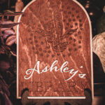 A laser cut and engraved sign made of 1/8" birch plywood stained dark walnut engraved with a woodcut of a A wooden arched library sign from The Griffin and Stag featuring an engraved dragon within a circle of stars, with "Ashley's Library" in white script and serif lettering.