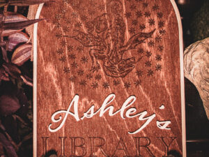 A laser cut and engraved sign made of 1/8" birch plywood stained dark walnut engraved with a woodcut of a A wooden arched library sign from The Griffin and Stag featuring an engraved dragon within a circle of stars, with "Ashley's Library" in white script and serif lettering.