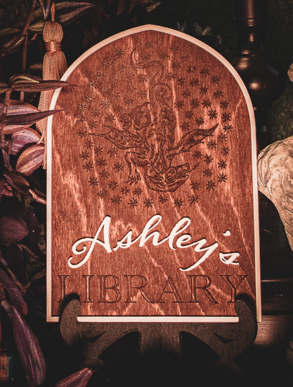 A laser cut and engraved sign made of 1/8" birch plywood stained dark walnut engraved with a woodcut of a A wooden arched library sign from The Griffin and Stag featuring an engraved dragon within a circle of stars, with "Ashley's Library" in white script and serif lettering.