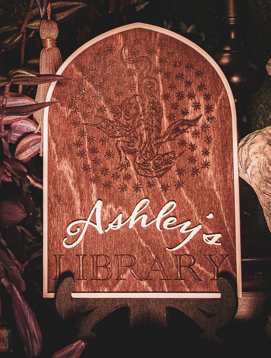 A laser cut and engraved sign made of 1/8" birch plywood stained dark walnut engraved with a woodcut of a medieval dragon surrounded by stars that reads, "Ashley's Library" where "Ashley" is painted white and a white frame resting on a laser cut stand made of 1/8" birch plywood stained ebony sitting on a bookshelf.