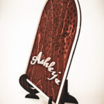 A wooden arched library sign from The Griffin and Stag featuring an engraved dragon within a circle of stars, with "Ashley's Library" in white script and serif lettering.