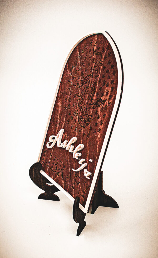 A wooden arched library sign from The Griffin and Stag featuring an engraved dragon within a circle of stars, with "Ashley's Library" in white script and serif lettering.