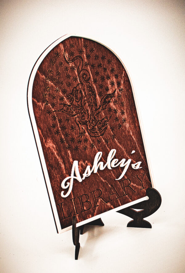 A wooden arched library sign from The Griffin and Stag featuring an engraved dragon within a circle of stars, with "Ashley's Library" in white script and serif lettering.
