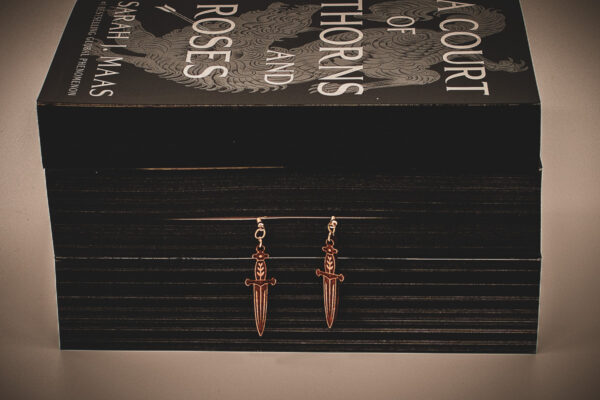 Small wooden dagger earrings with intricate floral hilt engravings, styled on a black-edged copy of A Court of Thorns and Roses by Sarah J. Maas.