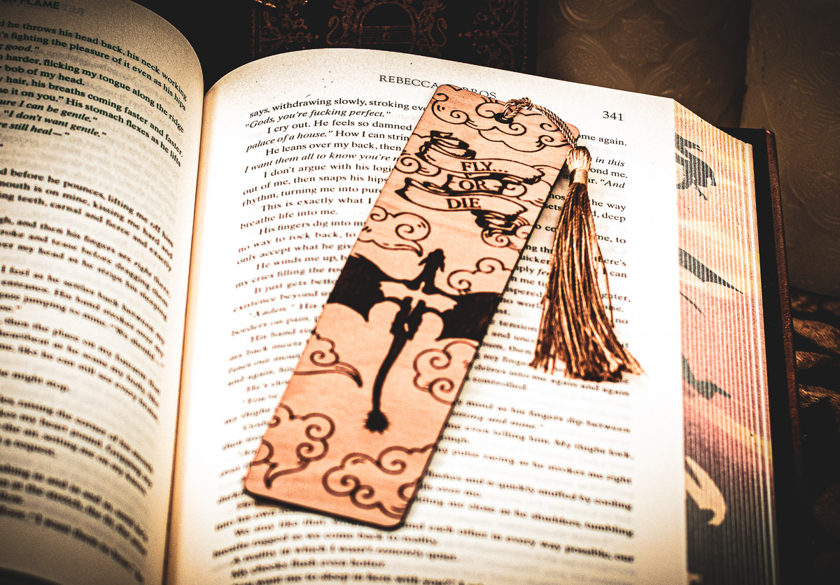 The Flight or Fate Marker - The Griffin and Stag Laser cut and engraved bookmark featuring two dragons in flight among clouds with a scroll of text that says "Fly or Die" on the page of an open book.
