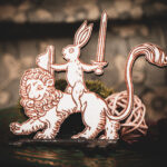 A laser cut and engraved magnet made of raw birch plywood in the shape of a rabbit holding a sword and shield riding on a lion leaning against a mossy rock on the cover of a book.