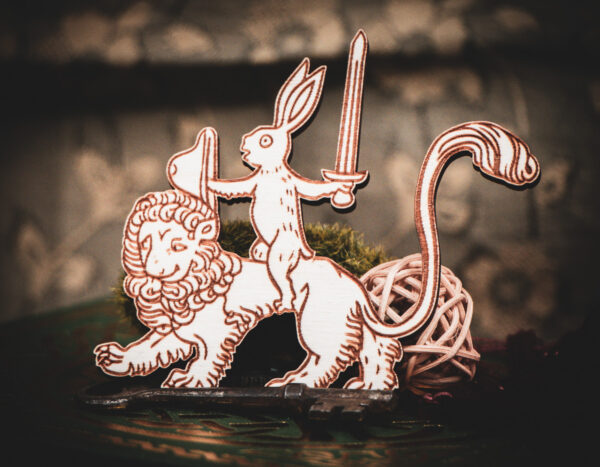 A laser cut and engraved magnet made of raw birch plywood in the shape of a rabbit holding a sword and shield riding on a lion leaning against a mossy rock on the cover of a book.