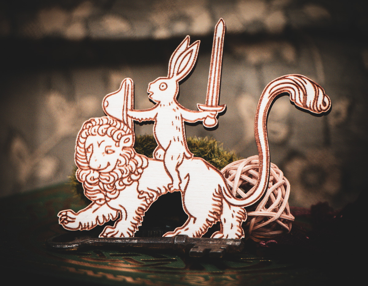 A laser cut and engraved magnet made of raw birch plywood in the shape of a rabbit holding a sword and shield riding on a lion leaning against a mossy rock on the cover of a book.