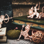 A group of three laser cut and engraved magnets made with raw birch plywood in the shapes of a snail archer, a satyr, and a rabbit riding a lion leaning among books, a hand sculpture, and mossy rocks.