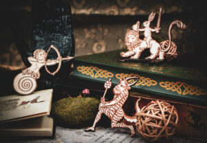 A group of three laser cut and engraved magnets made with raw birch plywood in the shapes of a snail archer, a satyr, and a rabbit riding a lion leaning among books, a hand sculpture, and mossy rocks.