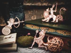 A group of three laser cut and engraved magnets made with raw birch plywood in the shapes of a snail archer, a satyr, and a rabbit riding a lion leaning among books, a hand sculpture, and mossy rocks.