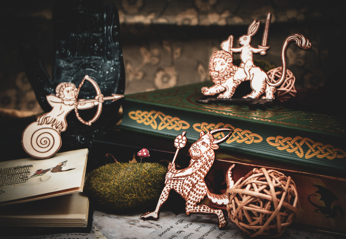The Illuminated Hearth - The Griffin and Stag A group of three laser cut and engraved magnets made with raw birch plywood in the shapes of a snail archer, a satyr, and a rabbit riding a lion leaning among books, a hand sculpture, and mossy rocks.