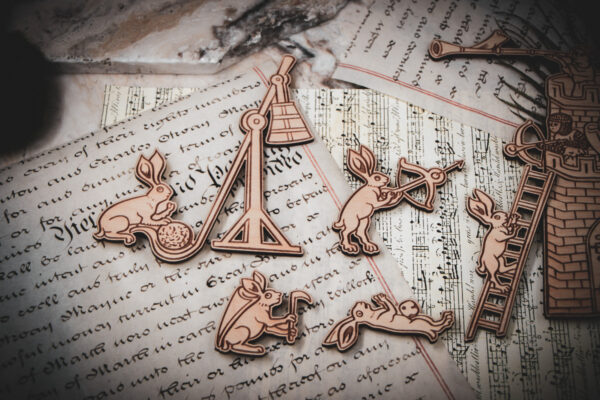 A close-up of four of a set of six laser cut and engraved birch magnets featuring rabbits waging battle against knights in a castle tower on top of various papers and a stone surface.