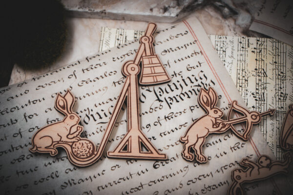 A close-up of two of a set of six laser cut and engraved birch magnets featuring rabbits waging battle against knights in a castle tower on top of various papers and a stone surface.