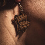A close-up profile of a person wearing a Renaissance Tome earring, highlighting the delicate laser-engraved lines of the open book pages against natural sunlight.
