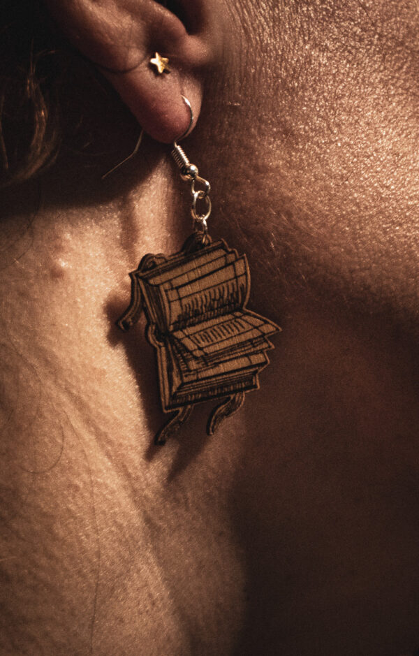 A close-up profile of a person wearing a Renaissance Tome earring, highlighting the delicate laser-engraved lines of the open book pages against natural sunlight.