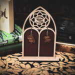 A pair of laser-cut wooden earrings, the "Litany of Lens," featuring detailed engravings of open Renaissance tomes with fluttering pages, displayed on a cathedral-style wooden stand with The Griffin and Stag logo.
