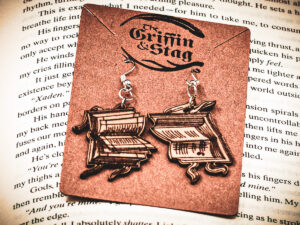 Handcrafted birch plywood book earrings styled on a brown "The Griffin and Stag" backing card, placed over an open copy of the fantasy novel Iron Flame.