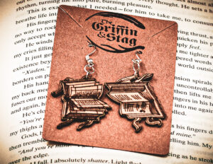 Handcrafted birch plywood book earrings styled on a brown "The Griffin and Stag" backing card, placed over an open copy of the fantasy novel Iron Flame.