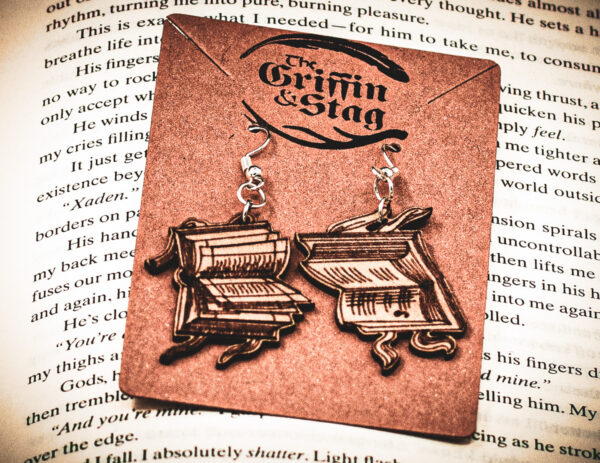 Handcrafted birch plywood book earrings styled on a brown "The Griffin and Stag" backing card, placed over an open copy of the fantasy novel Iron Flame.