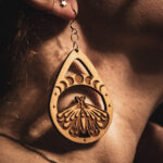 A close-up side profile of a person wearing a Lunar Metamorphosis earring, showcasing the detailed engraving and natural wood grain against a warm, sunlit background.