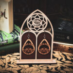 A pair of teardrop-shaped wooden earrings, the "Lunar Metamorphosis," featuring a laser-cut moth beneath a series of moon phases, displayed on a custom gothic-arch stand with The Griffin and Stag logo.