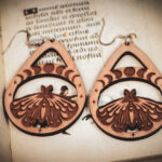 Handcrafted birch wood moth earrings with moon phase engravings and star accents, styled over an antique manuscript featuring medieval marginalia.