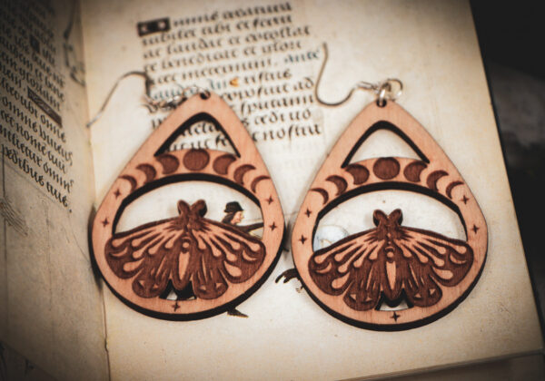 Handcrafted birch wood moth earrings with moon phase engravings and star accents, styled over an antique manuscript featuring medieval marginalia.