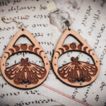 Two laser cut and engraved earrings featuring a luna moth with a band overhead showing the phases of the moon within a teardrop frame on top of handwritten pages of calligraphy.