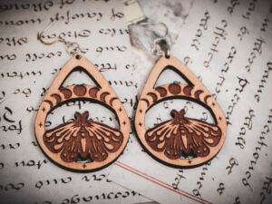 Two laser cut and engraved earrings featuring a luna moth with a band overhead showing the phases of the moon within a teardrop frame on top of handwritten pages of calligraphy.