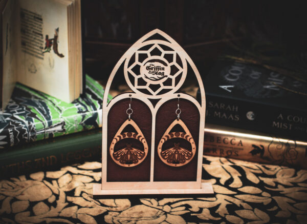 A pair of teardrop-shaped wooden earrings, the "Lunar Metamorphosis," featuring a laser-cut moth beneath a series of moon phases, displayed on a custom gothic-arch stand with The Griffin and Stag logo.