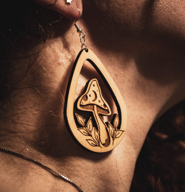 A close-up side profile of a person wearing a Mycelial Mystic earring, showcasing the detailed engraving and natural wood grain against a warm, sunlit background.
