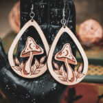 The teardrop mushroom earrings styled on a black, ornate palmistry hand model, surrounded by dark academia-themed decor and vintage books.