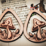 Handcrafted birch wood mushroom earrings with celestial engravings and leaf accents, styled over an antique manuscript featuring medieval marginalia.