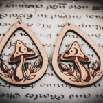 Two laser cut and engraved earrings featuring a mushroom with a crescent moon and stars on the cap surrounded by leaves within a teardrop frame on top of a paper with calligraphy.