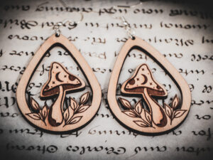 Two laser cut and engraved earrings featuring a mushroom with a crescent moon and stars on the cap surrounded by leaves within a teardrop frame on top of a paper with calligraphy.