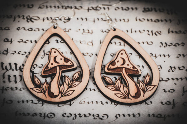 Two laser cut and engraved earrings featuring a mushroom with a crescent moon and stars on the cap surrounded by leaves within a teardrop frame on top of a paper with calligraphy.