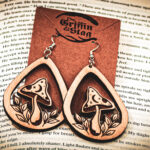 Two laser cut and engraved earrings featuring a mushroom with a crescent moon and stars on the cap surrounded by leaves within a teardrop frame hanging from a kraft paper card on top of the page of an open book.