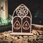 A pair of teardrop-shaped wooden earrings, the "Mycelial Mystic," featuring a laser-cut mushroom with a crescent moon and star detail on the cap, displayed on a custom gothic-arch stand with The Griffin and Stag logo.