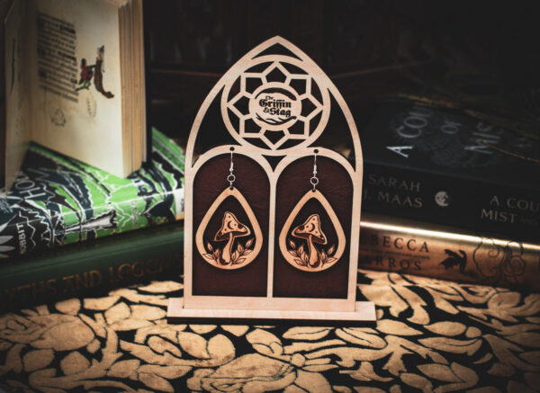A pair of teardrop-shaped wooden earrings, the "Mycelial Mystic," featuring a laser-cut mushroom with a crescent moon and star detail on the cap, displayed on a custom gothic-arch stand with The Griffin and Stag logo.