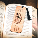 A laser-engraved wooden bookmark featuring a mountain range under stars with the quote "To the stars who listen... and the dreams that are answered," resting on an open fantasy novel.